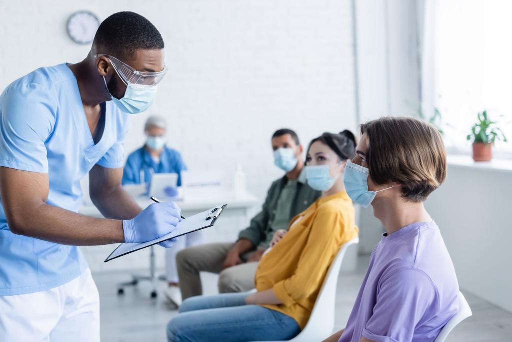 médecin afro-américain en masque médical écrit sur presse-papiers près des patients dans le centre de vaccination - Photo, image