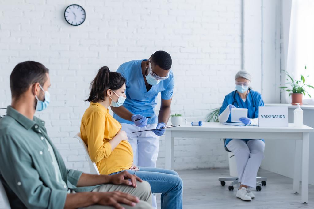 Afrikanisch-amerikanischer Arzt hält Klemmbrett und Stift in der Nähe einer Schwangeren in einem Impfzentrum - Foto, Bild