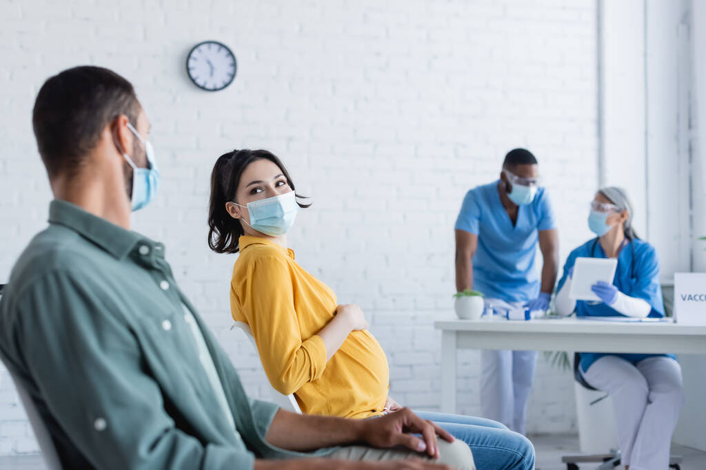 mujer embarazada con máscara médica cerca del hombre y los médicos interracial borrosa en el centro de vacunación - Foto, imagen