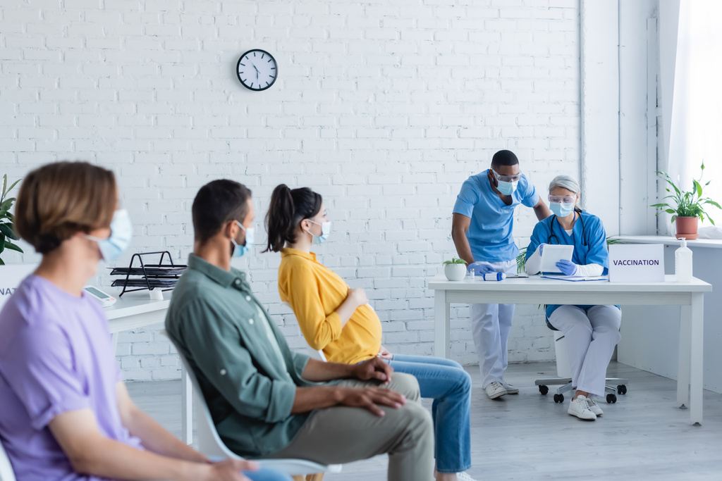 hamile kadın ve maskeli genç erkekler aşı merkezinde çok ırklı doktorların yanında oturuyorlar. - Fotoğraf, Görsel