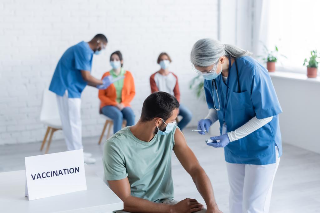 Afro-Amerikan meslektaşı hastalarla konuşurken tıbbi maskeli adamların yanında aşı tutan olgun bir Asyalı hemşire.  - Fotoğraf, Görsel