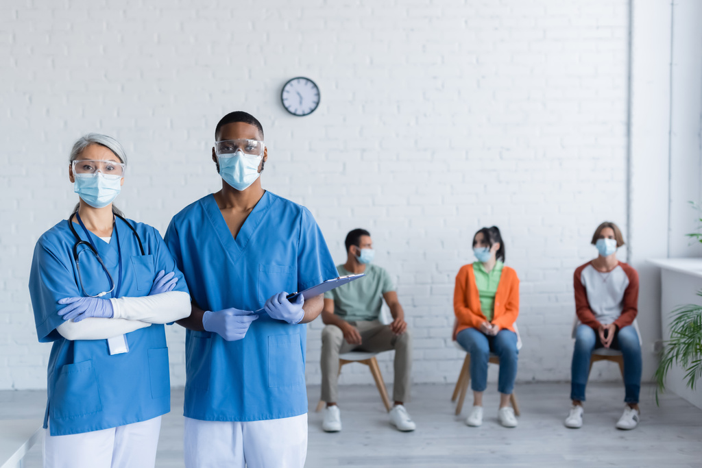medici interrazziale in maschere mediche e occhiali guardando la fotocamera vicino ai pazienti su sfondo sfocato - Foto, immagini