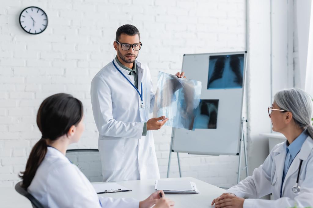 lekarz w okularach pokazujący prześwietlenie płuc wielonarodowym kolegom podczas rady lekarskiej - Zdjęcie, obraz
