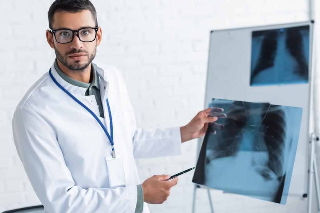 nuori lääkäri silmälaseissa osoittaa keuhkojen röntgeniin katsoessaan kameraa - Valokuva, kuva