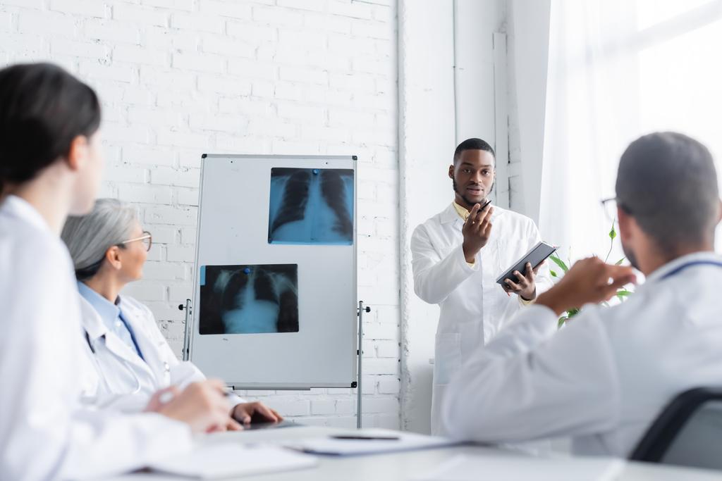 афроамериканський лікар жестикулює, розмовляючи з розмитими колегами поблизу фліп-чарту з рентгенівськими променями легень
 - Фото, зображення