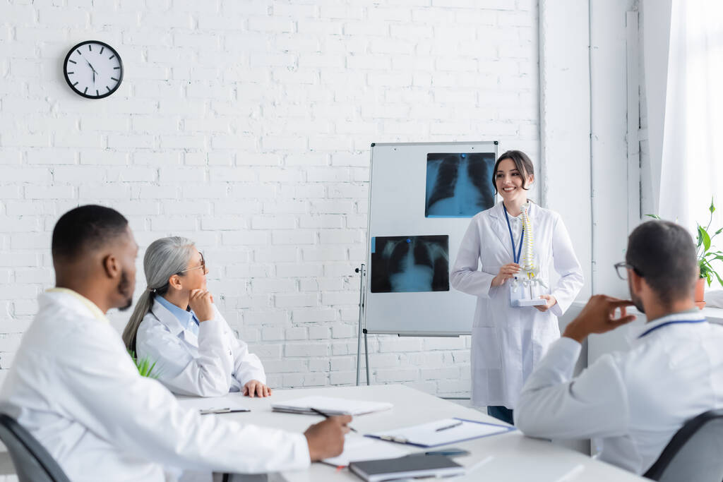 hymyilevä lääkäri seisoo lähellä flip kaavio keuhkojen röntgenkuvat ja osoittaa selkärangan malli monikansallisille kollegoille  - Valokuva, kuva