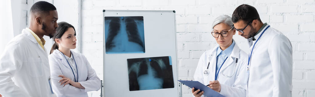 mature asiatique médecin pointant vers presse-papiers près de flip chart avec poumons rayons X et jeunes collègues multiethniques, bannière - Photo, image
