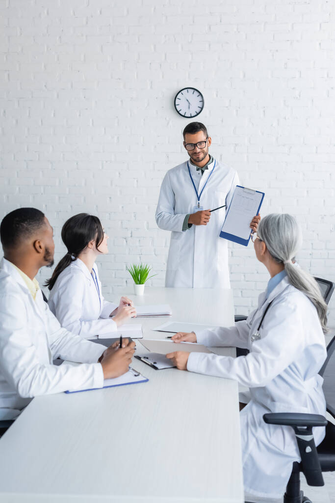 uśmiechnięty lekarz w okularach wskazujący na schowek w pobliżu wielonarodowych kolegów siedzących przy biurku - Zdjęcie, obraz