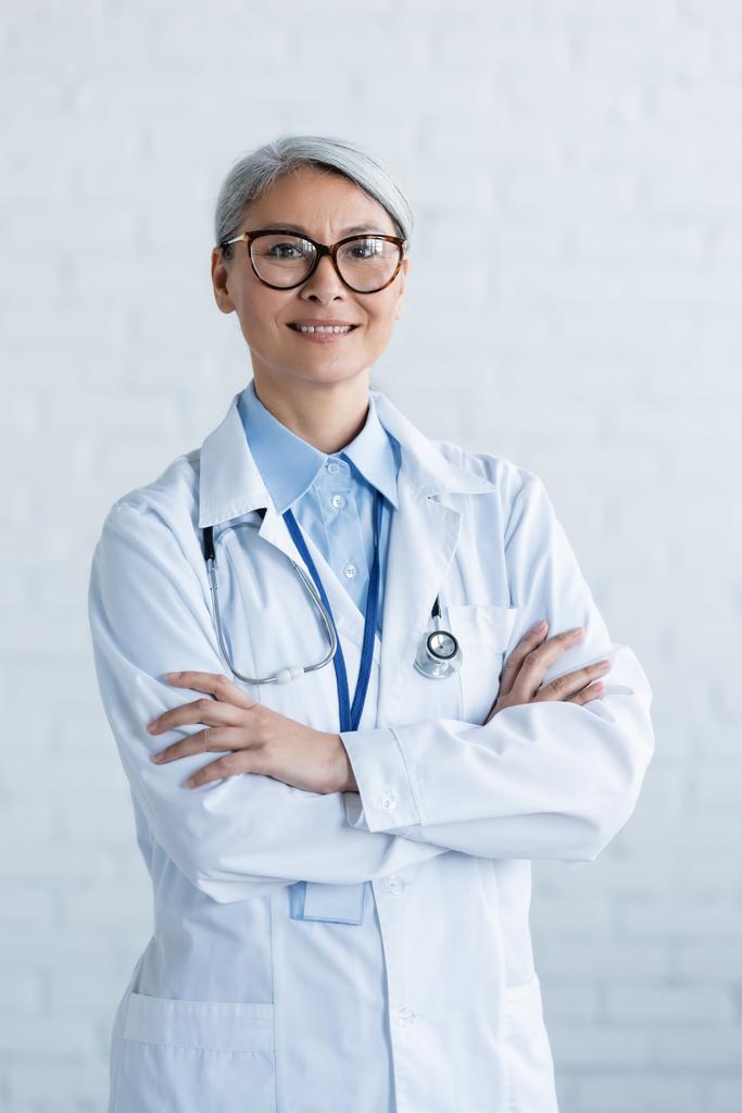 maduro asiático médico en blanco abrigo y gafas de pie con los brazos cruzados y sonriendo a la cámara en blanco - Foto, imagen