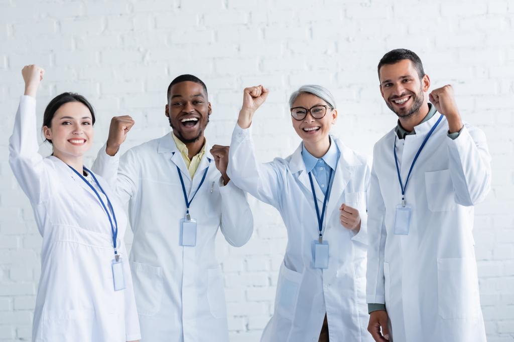 Aufgeregte multikulturelle Ärzte in weißen Kitteln mit Namensschildern, die Jubelgeste zeigen - Foto, Bild