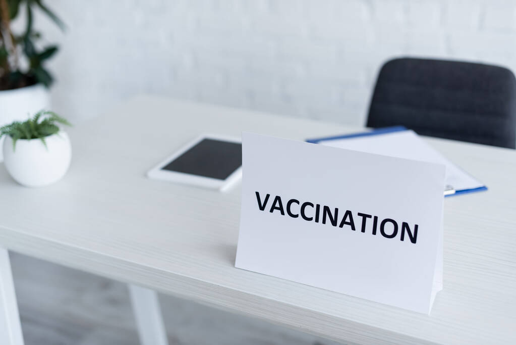 placa con letras de vacunación cerca de la tableta digital borrosa en el escritorio en la clínica - Foto, Imagen
