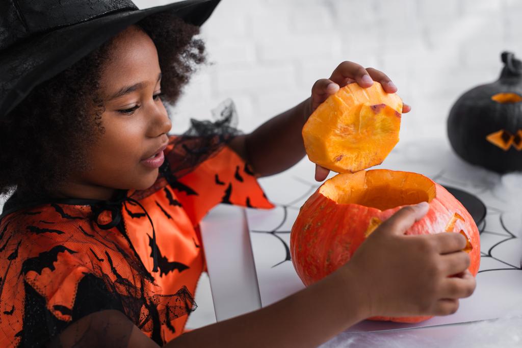 アフリカ系アメリカ人の少女魔女ハロウィンの衣装でカボチャを彫る - 写真・画像