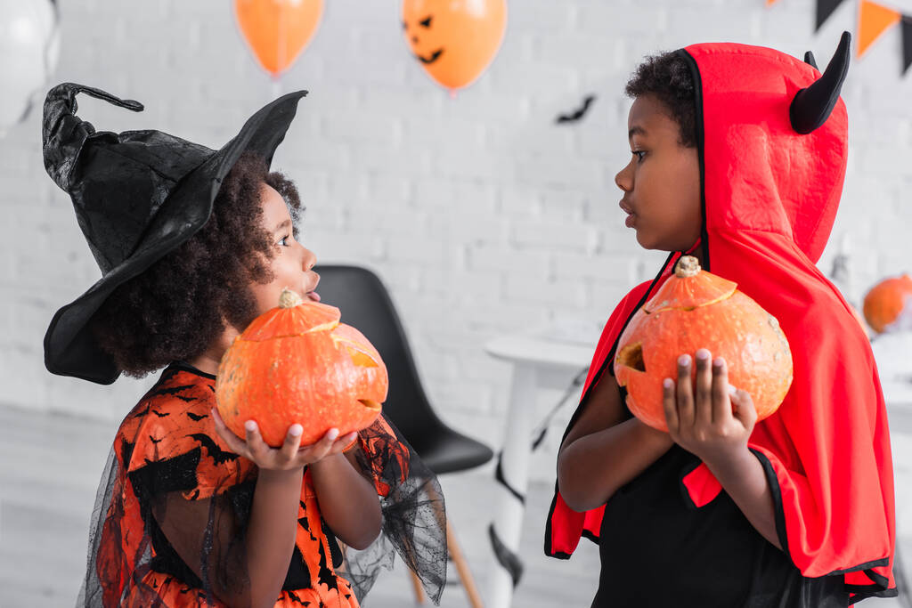 zijaanzicht van verrast Afrikaans-Amerikaanse kinderen in halloween kostuums houden gesneden pompoenen thuis - Foto, afbeelding