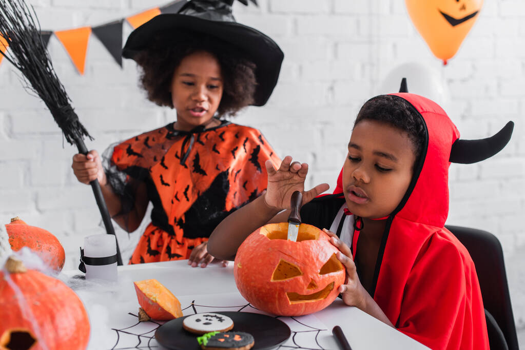 africano americano ragazzo in halloween costume scultura zucca vicino sfocata sorella con scopa  - Foto, immagini