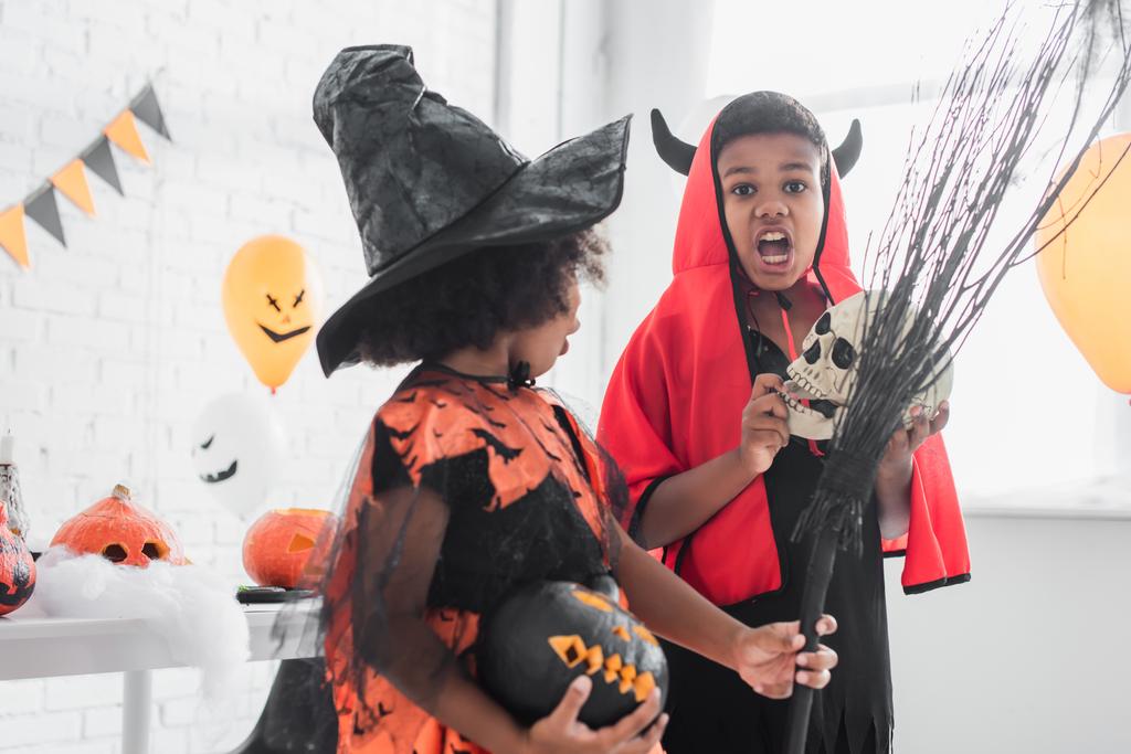 effrayant garçon afro-américain en costume d'Halloween tenant crâne près de sœur avec balai et citrouille sculptée  - Photo, image