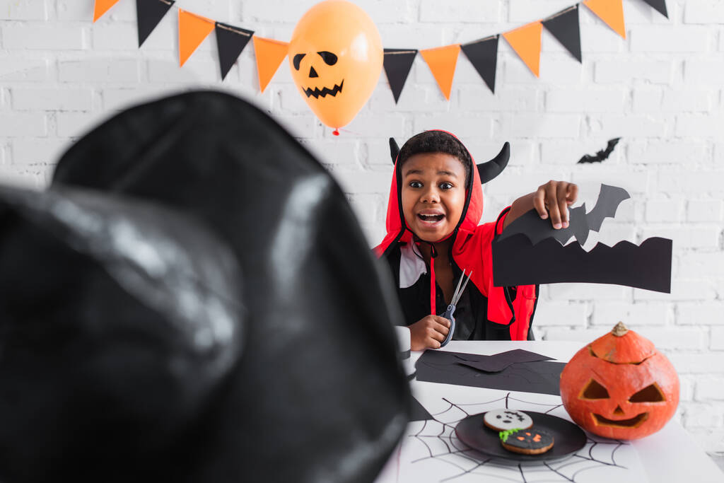 opgewonden Afrikaans amerikaanse jongen in halloween kostuum tonen vleermuis template te wazig zus in puntige hoed  - Foto, afbeelding