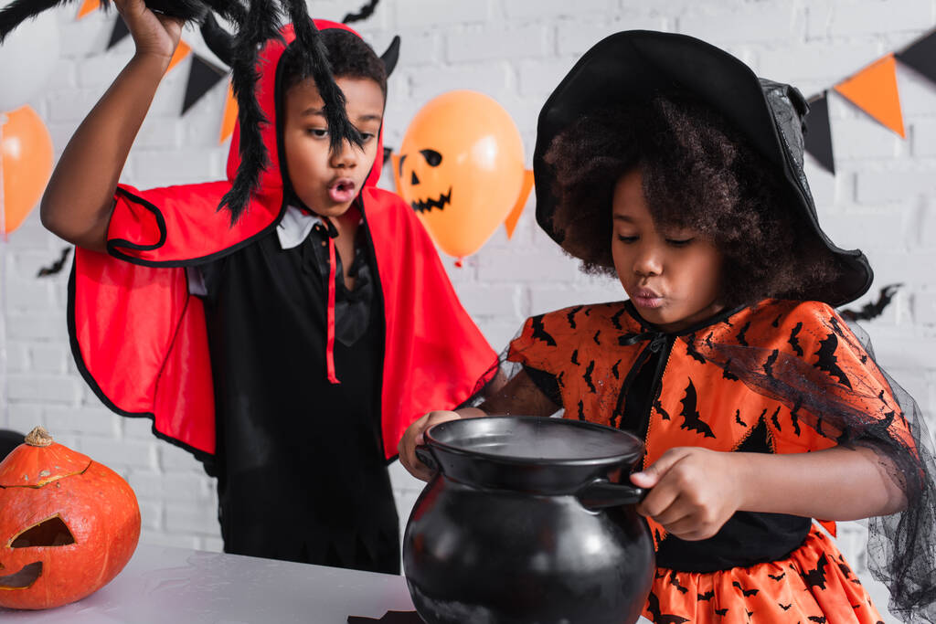 africano americano ragazza in halloween costume tenendo strega calderone con pozione vicino fratello con giocattolo ragno - Foto, immagini