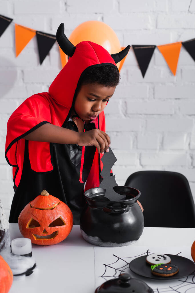 Afryki amerykański chłopiec w diabeł halloween kostium trzymając papier wyciąć nietoperz powyżej kotła - Zdjęcie, obraz