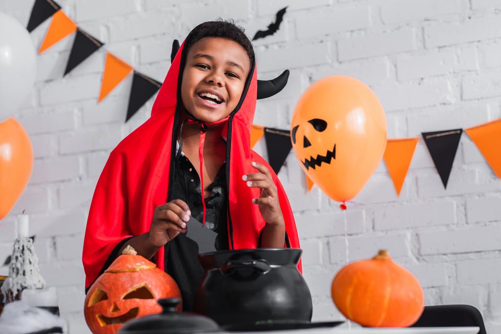 happy african american boy in devil halloween costume holding paper cut bat above cauldron - Photo, Image