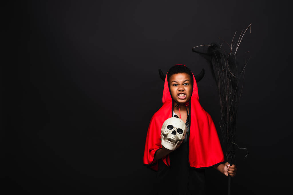 Θυμωμένο αφροαμερικανό αγόρι που κρατάει σκούπα και κρανίο ενώ χαμογελάει απομονωμένο στα μαύρα - Φωτογραφία, εικόνα