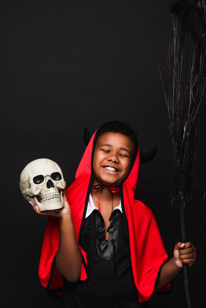 Χαρούμενο Αφροαμερικανό αγόρι που κρατάει σκούπα και κρανίο απομονωμένο σε μαύρο.  - Φωτογραφία, εικόνα