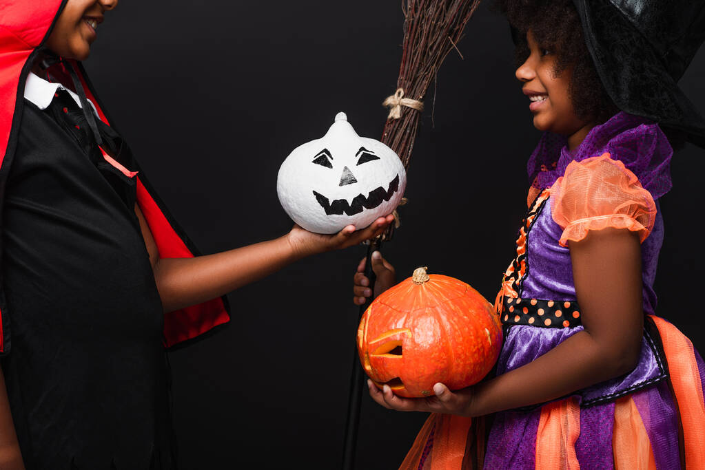 sorrindo crianças afro-americanas em trajes halloween segurando abóboras isoladas em preto  - Foto, Imagem
