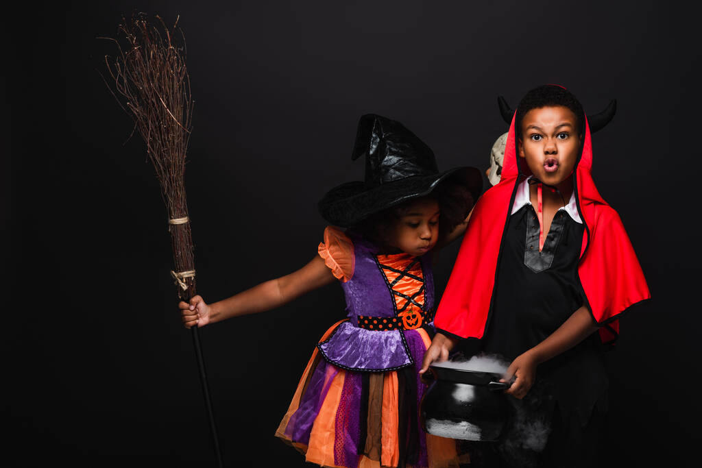 Afrikaans amerikaans meisje in halloween kostuum houden bezem en blazen op ketel met toverdrank in de buurt broer geïsoleerd op zwart  - Foto, afbeelding