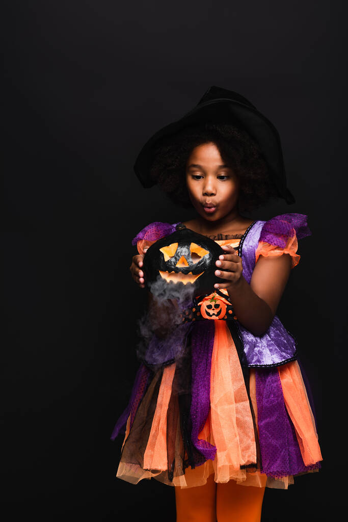 Αφροαμερικανή κοπέλα με αποκριάτικη στολή που κοιτάζει σκαλιστή κολοκύθα με καπνό που απομονώνεται στο μαύρο  - Φωτογραφία, εικόνα