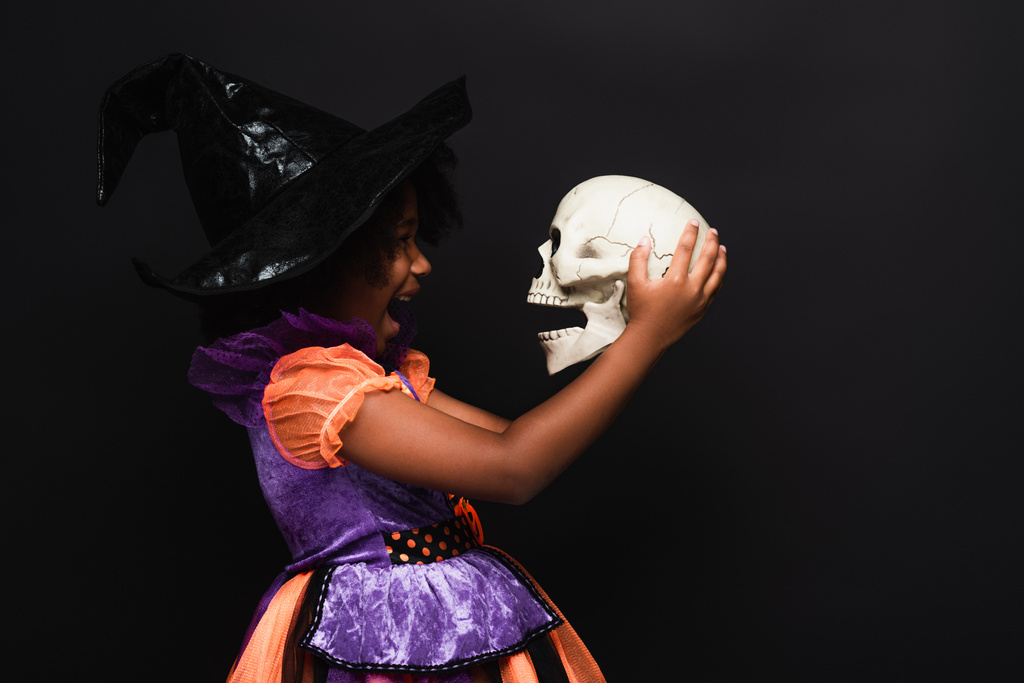 vue latérale de excitée afro-américaine fille en costume de sorcière halloween tenant crâne isolé sur noir - Photo, image
