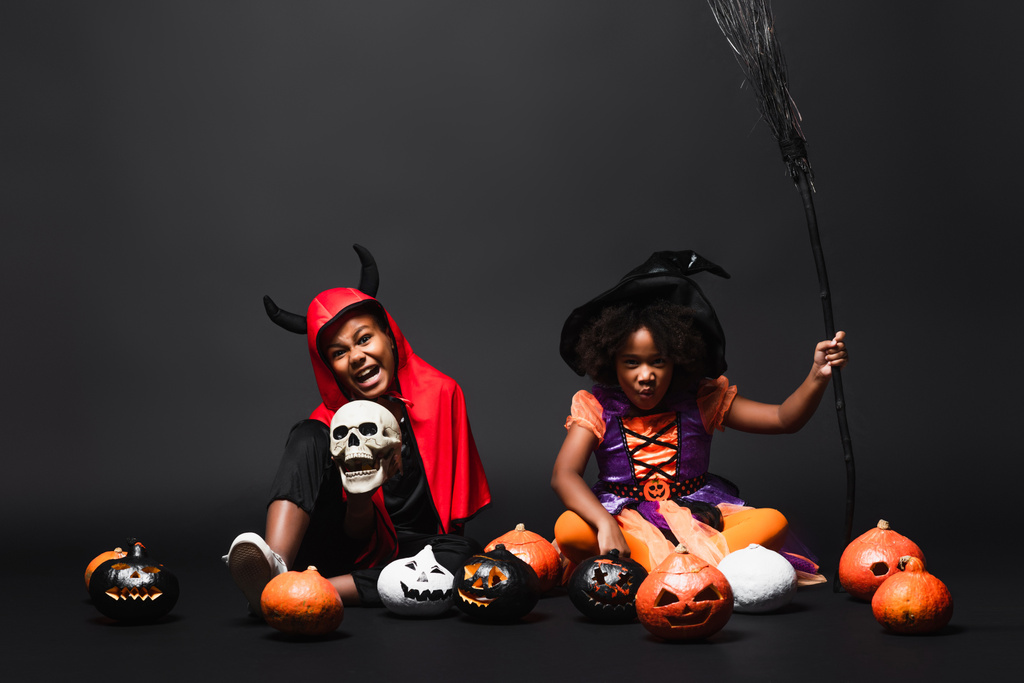 Afrikanisch-amerikanische Kinder in Halloween-Kostümen mit Totenkopf und Besen in der Nähe von Kürbissen auf schwarz  - Foto, Bild