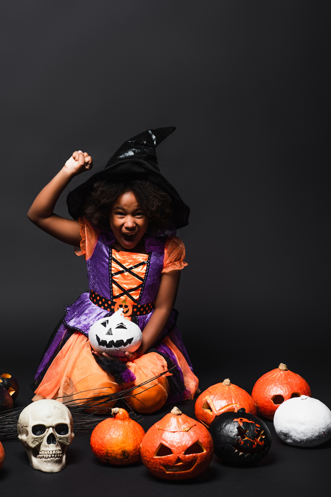 excited african american child in witch costume and pointed hat near carved pumpkins on black - Photo, Image