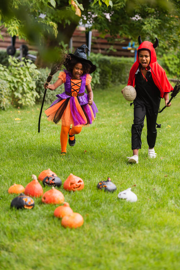 eccitati bambini africani americani in costumi di Halloween che tengono scope e corrono sul cortile  - Foto, immagini