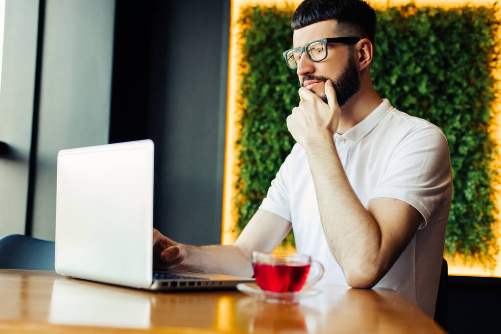 Portré egy jóképű, sikeres férfiról, aki teát iszik és laptopot néz egy kávézóban ülve, üzletember reggelizik egy kávézóban és laptopot használ, az interneten böngészve - Fotó, kép