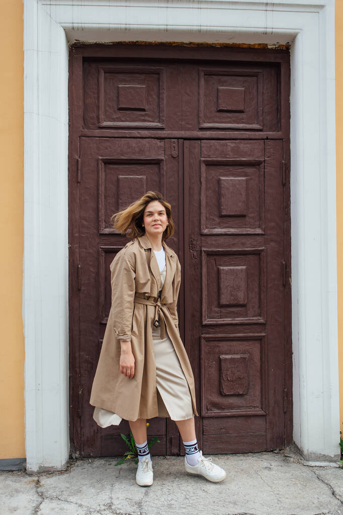 Joven mujer milenaria sonriente con pelo salvaje vestida con un abrigo de otoño posando cerca de la puerta de un viejo edificio. - Foto, imagen