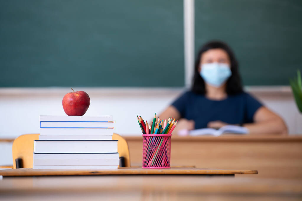 Επιστροφή στο σχολείο Concept. Μήλο, αξεσουάρ και βιβλία στην τάξη, δάσκαλος φορώντας μάσκα στο παρασκήνιο. - Φωτογραφία, εικόνα