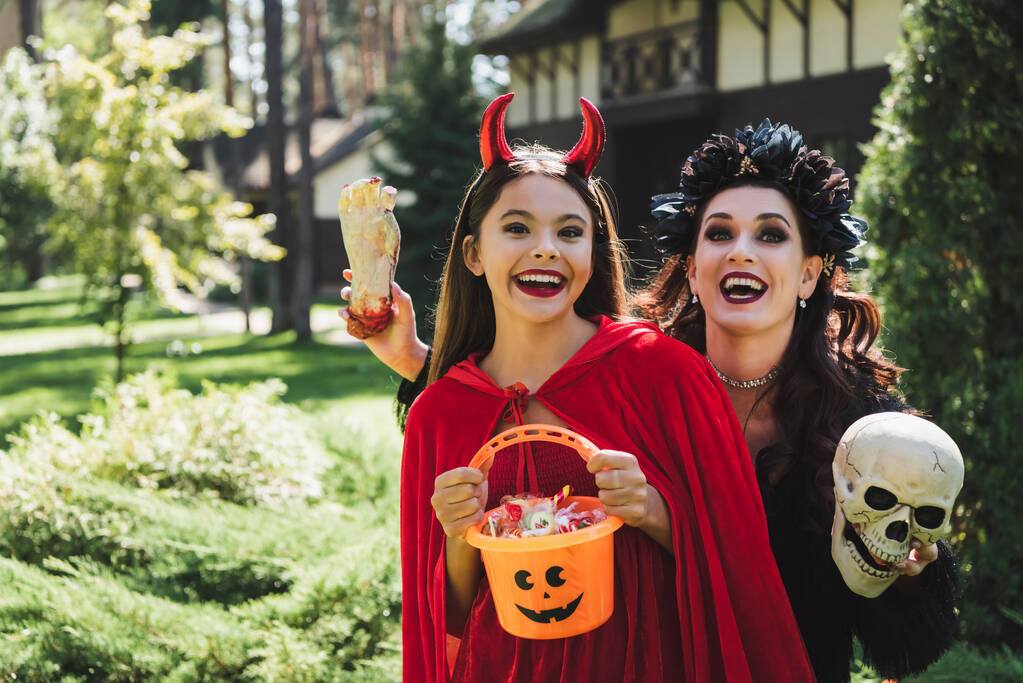 excitada mamá y su hija en trajes de Halloween diablo y vampiro, con cráneo, mano de juguete y cubo de caramelos - Foto, imagen