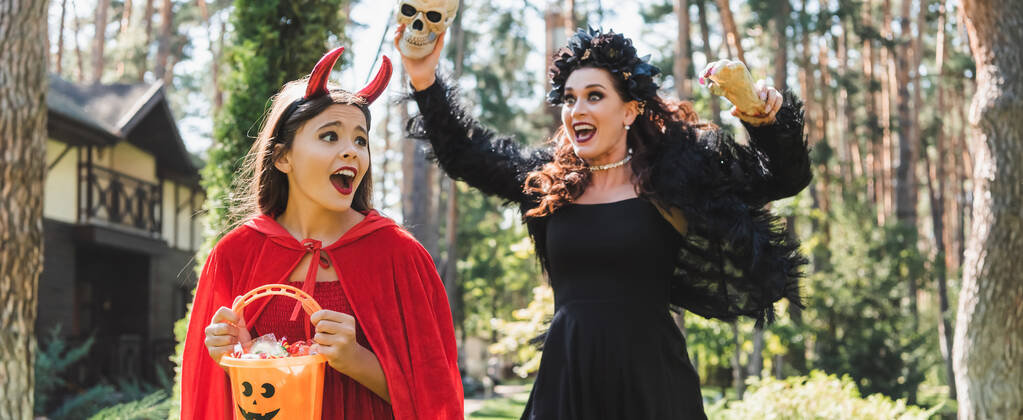 mujer en traje de halloween vampiro asustando a la hija con la mano de juguete y el cráneo, bandera - Foto, Imagen