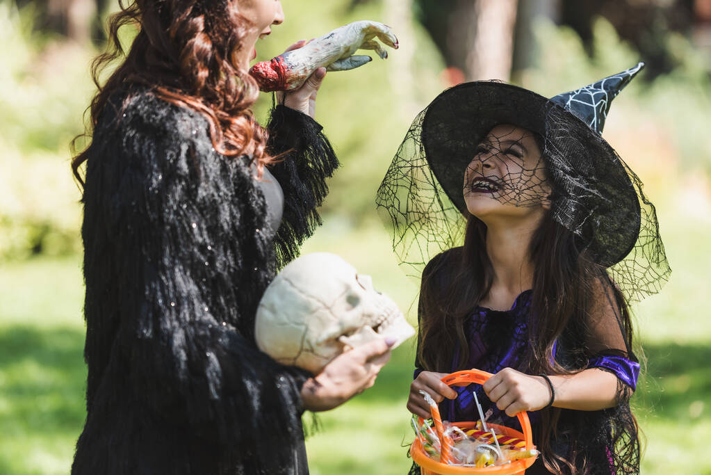 wazig vrouw in zwart halloween kostuum schrikken vrolijke dochter met speelgoed hand - Foto, afbeelding