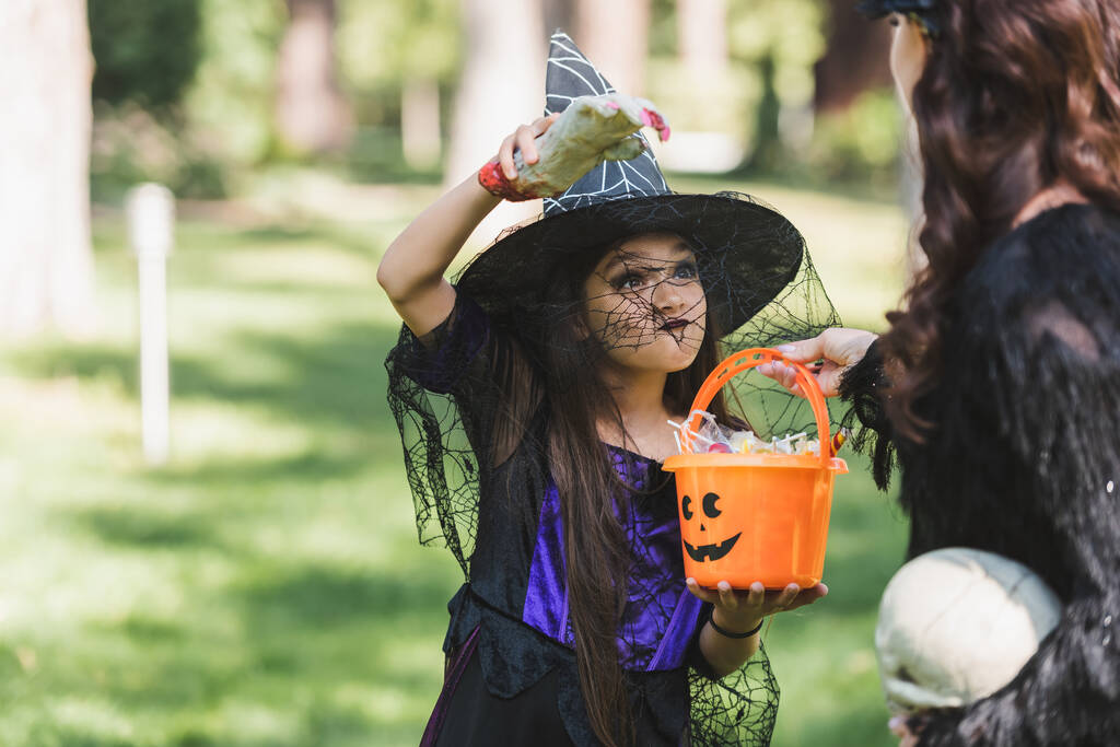 menina em chapéu de bruxa segurando balde com doces e assustador mãe com mão de brinquedo - Foto, Imagem