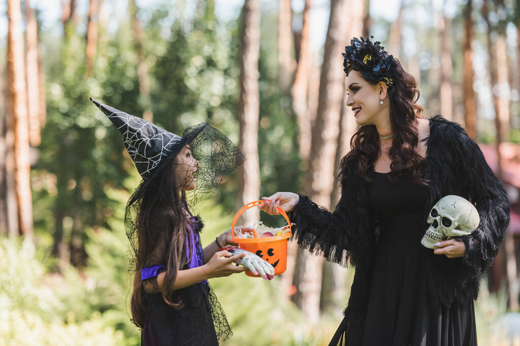 gelukkig moeder en dochter in Halloween kostuums, met emmer snoep en schedel in het bos - Foto, afbeelding
