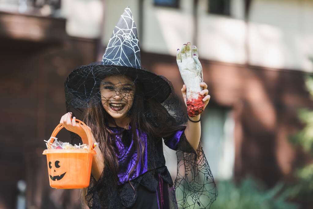 взволнованная девушка в костюме ведьмы на Хэллоуин, держа игрушечную руку и ведро со сладостями - Фото, изображение