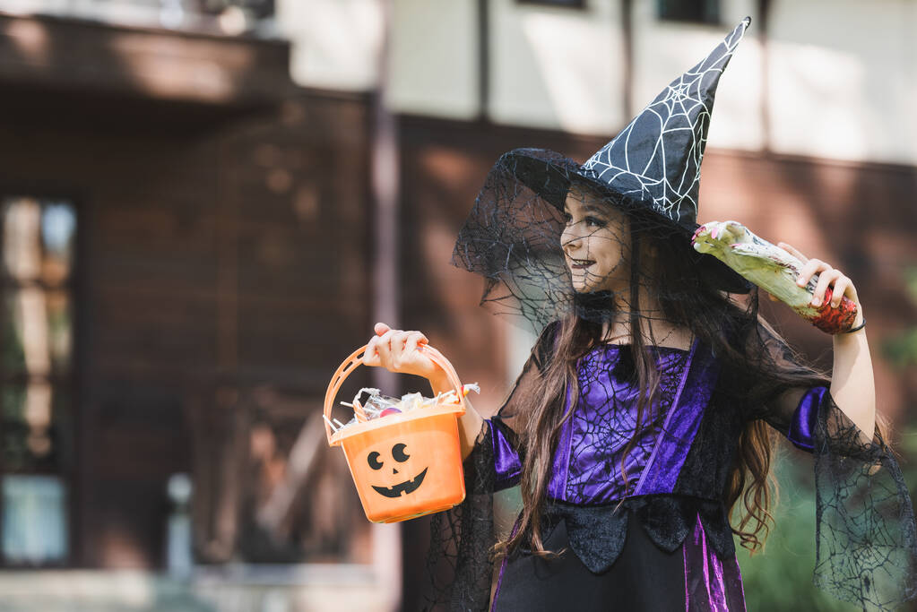魔女ハロウィンの衣装を着た幸せな子供おもちゃの手と屋外で見てキャンディー付きのバケツ - 写真・画像