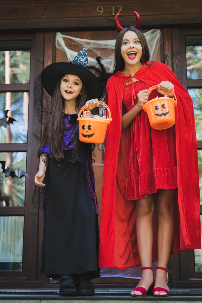 hermanas asombradas en trajes de bruja y diablo halloween, con cubos de dulces en el porche de la cabaña - Foto, imagen