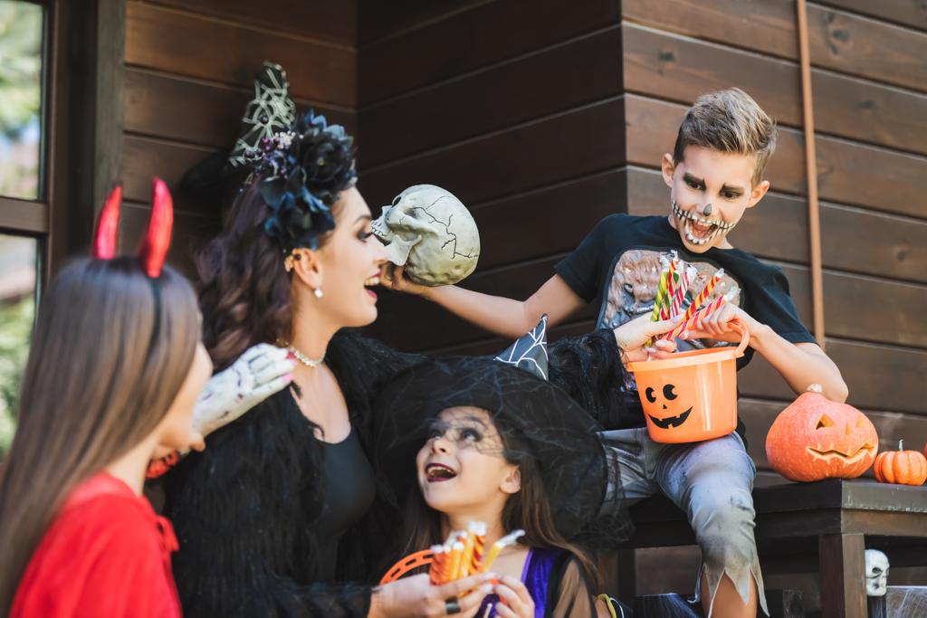 chlapec drží kbelík s bonbóny v blízkosti ohromený maminka a sestry v halloween kostýmy - Fotografie, Obrázek