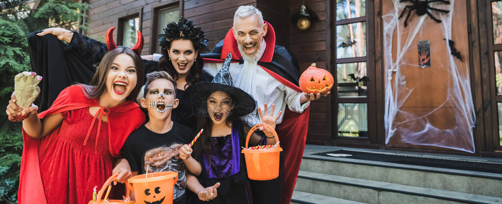 family in spooky costumes holding halloween attributes while growling at camera, banner - Photo, Image