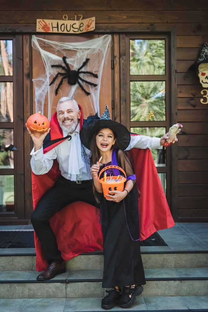 menina alegre e pai em trajes de Halloween sorrindo no alpendre da casa com decoração - Foto, Imagem
