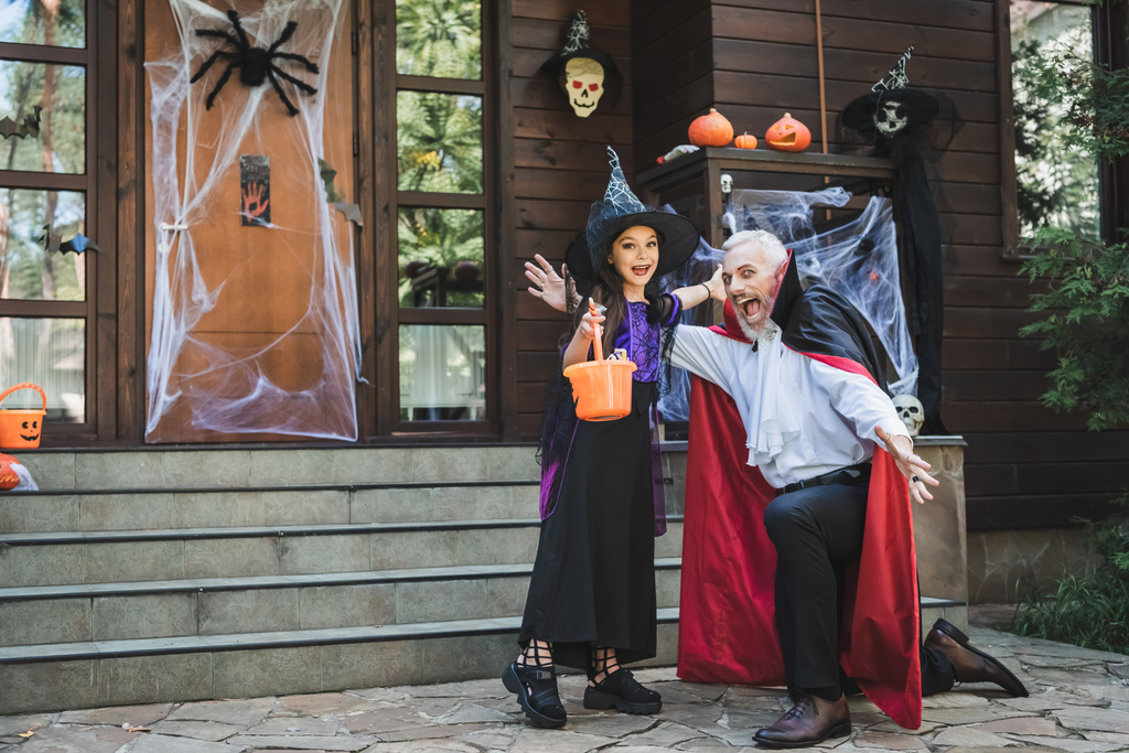 吸血鬼の衣装を着た興奮した男とハロウィンの装飾が施されたポーチ近くのお菓子のバケツを持つ女の子 - 写真・画像