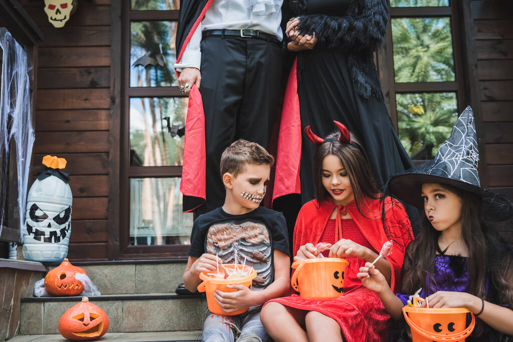ハロウィンの衣装を着た子供たちが親のそばにお菓子のバケツを持ってコテージの階段に座って - 写真・画像