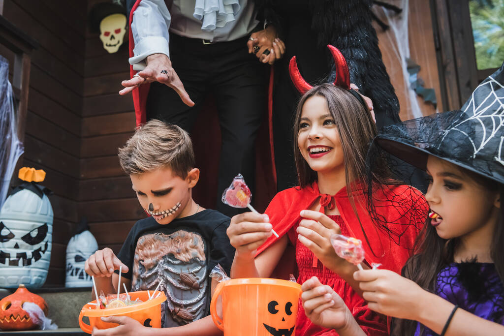 enfants heureux en costumes d'Halloween tenant des seaux avec sucettes près des parents - Photo, image