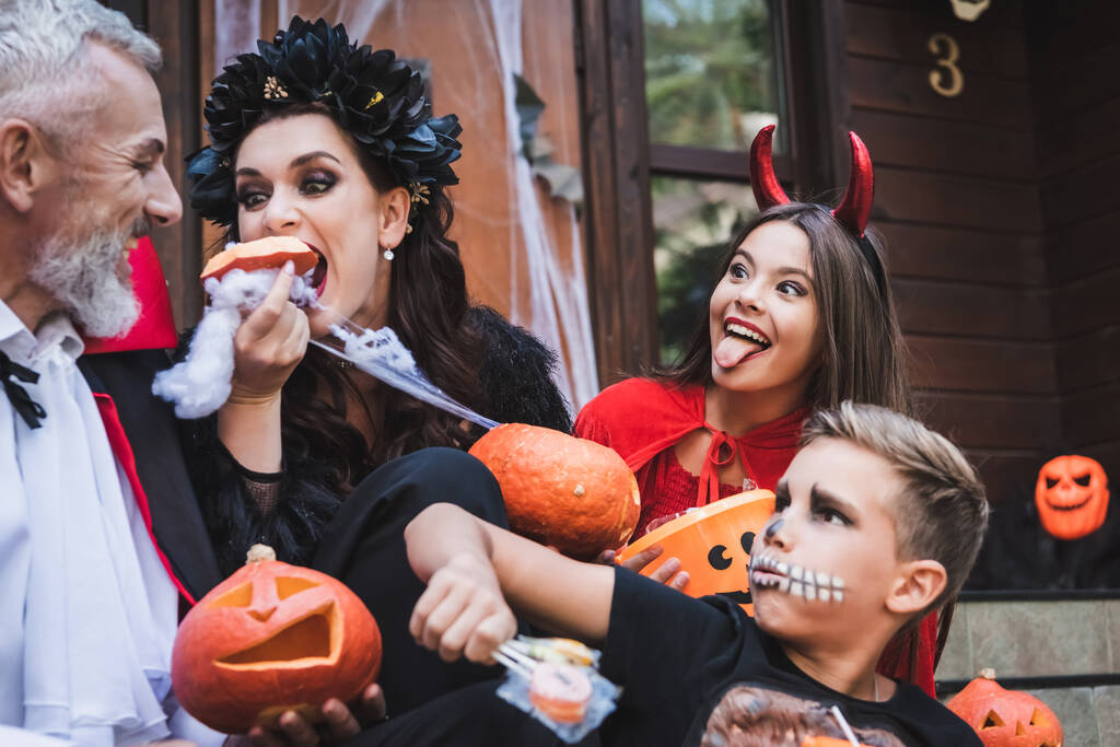 不気味なハロウィンの衣装で興奮した家族の近くで生のカボチャを食べる女性 - 写真・画像